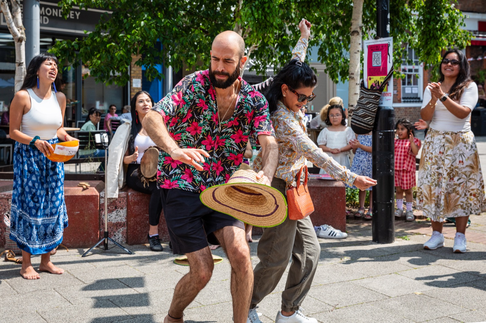 A community celebration in Bermondsey: people dancing in the street, families watching, a sunlit square.